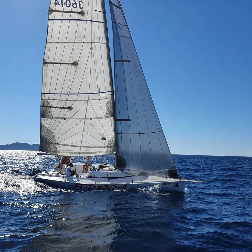 A sailing boat with a large sail glides on the blue waters under a clear sky, with several people on board. The boat is participating in a Marlin Sailing Academy sailing course near Zadar, Croatia.