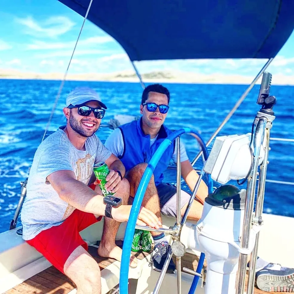 Two men sitting at the helm of a sailboat during Marlin Sailing Academy sailing course, holding drinks, under a blue canopy with the sea and distant land visible in the background.